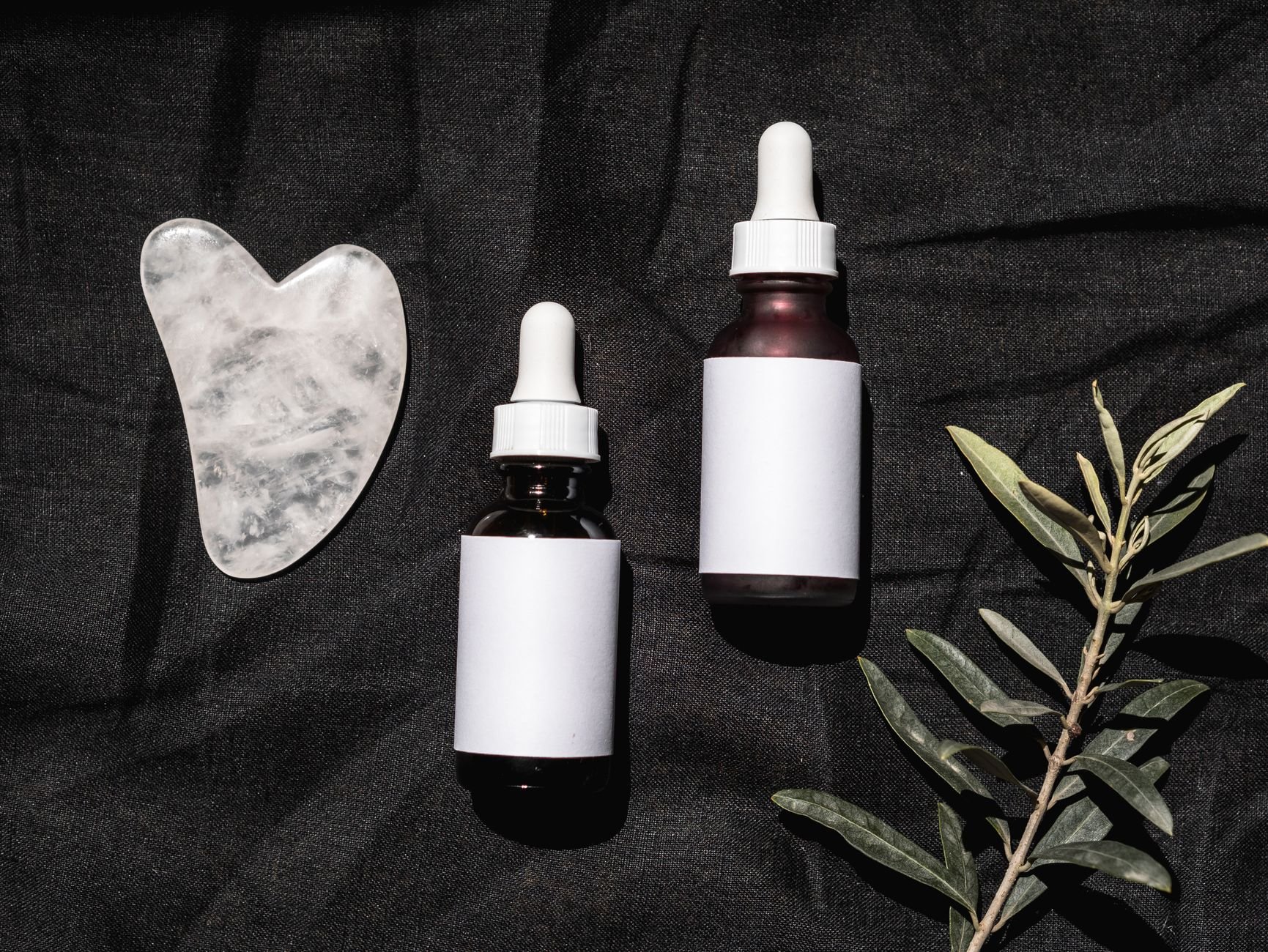 Minimalist flatlay of skincare bottles, gua sha, and botanicals on dark fabric
